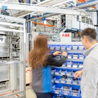 Two technicians in uniforms working at blue storage bins in an industrial facility with electrical panels.