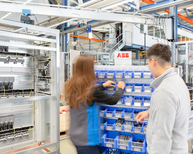 Two technicians in uniforms working at blue storage bins in an industrial facility with electrical panels.