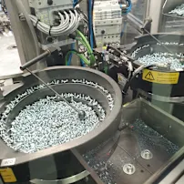 Máquina industrial con dos bandejas circulares llenas de tornillos plateados, rodeada de cables y paneles de control.