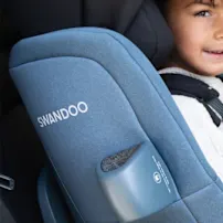 Kind in einem blauen Swandoo-Autositz, lächelnd, trägt ein weißes Hemd und Jeans, sitzt auf dem Rücksitz eines Autos.