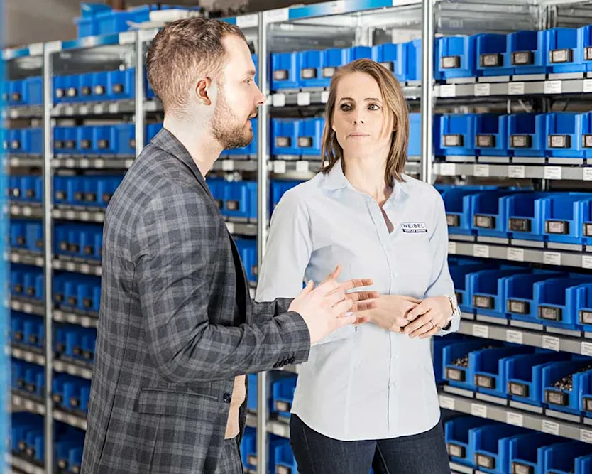 A man and a woman standing in front of a rack of blue Bossard Smartbins at Weibel