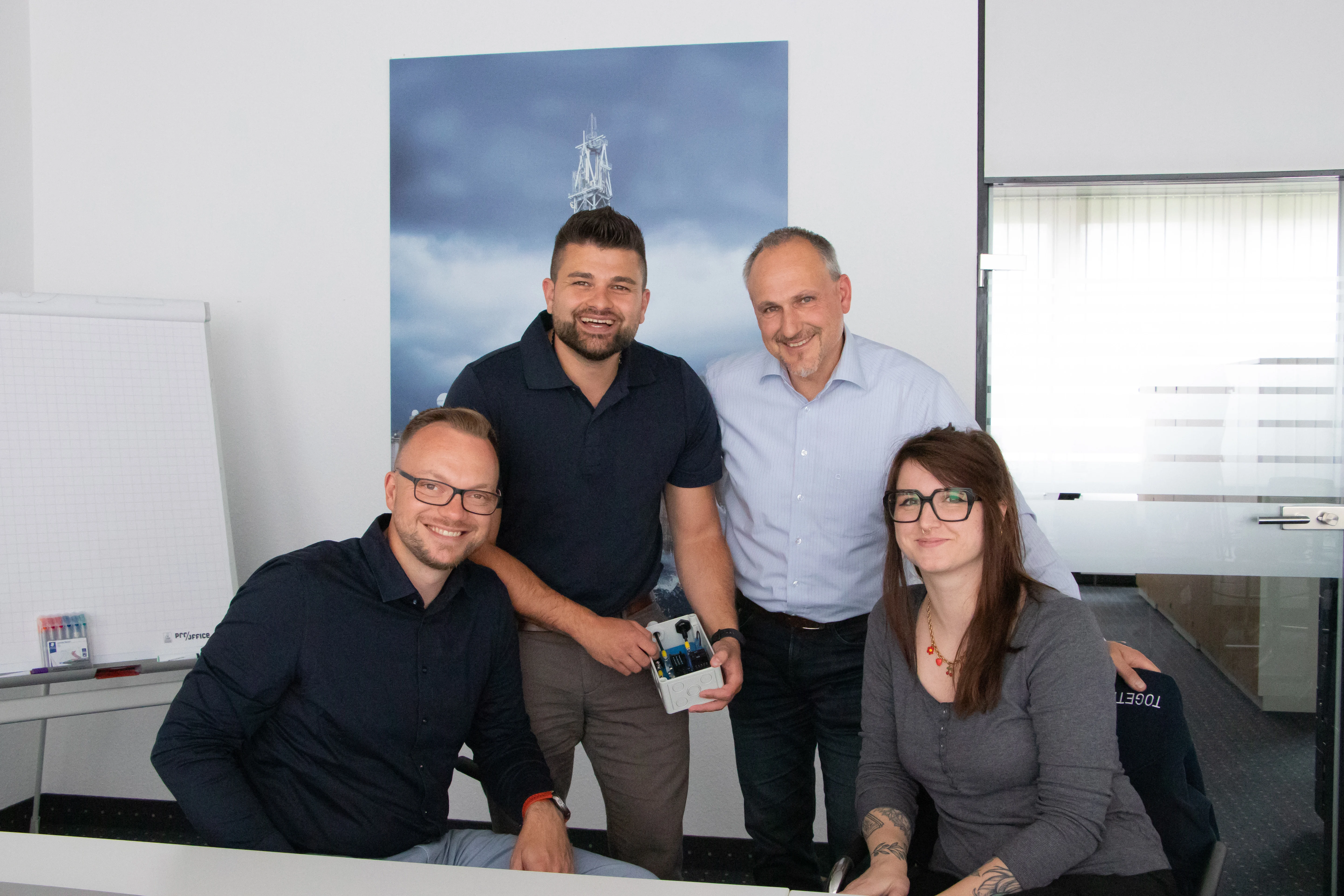 Vier Personen sitzen und stehen in einem Besprechungsraum, lächeln in die Kamera. Einer hält ein kleines Gehäuse mit Bauteilen. Im Hintergrund eine Glastüre, eine Staffelei und ein Poster mit einem Turm.