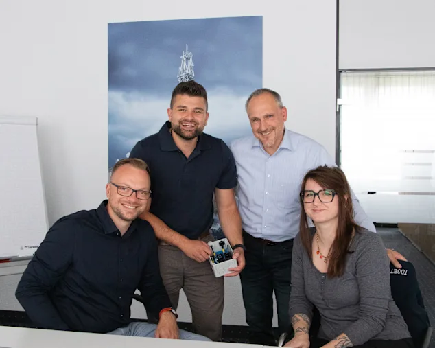 Vier Personen sitzen und stehen in einem Besprechungsraum, lächeln in die Kamera. Einer hält ein kleines Gehäuse mit Bauteilen. Im Hintergrund eine Glastüre, eine Staffelei und ein Poster mit einem Turm.
