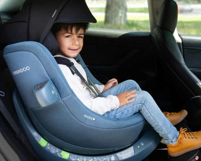 lächelndes Kind in einem Autositz mit Sicherheitsgurt, Marke SWANDOO sichtbar.