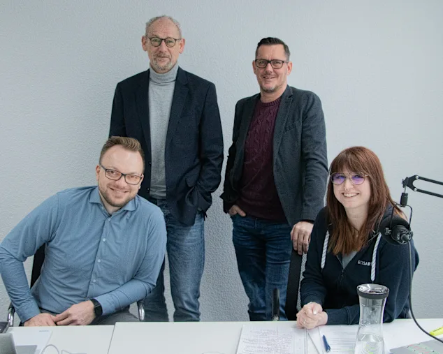 Vier Experten sitzen im Aufnahmestudio, weiße Wand im Hintergrund, Broadcast Mikrofone auf dem Tisch im Vordergrund