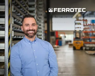 Ferrotec-Geschäftsführer Asim Aghdiri lehnt an einem metallfarbenem Lagerregal. Er trägt ein blaues Hemd und lächelt. Im Hintergrund eine Lagerhalle verschwommen sichtbar. Oben Rechts das Fettotec Logo in weiß.