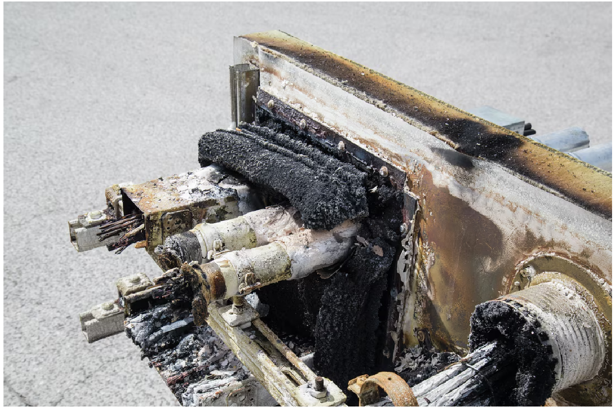 Close-up of a fire-tested cable penetration seal with charred cables, damaged firestop material, exposed conductors, and heat-discoloured metal, illustrating the effects of severe fire exposure.