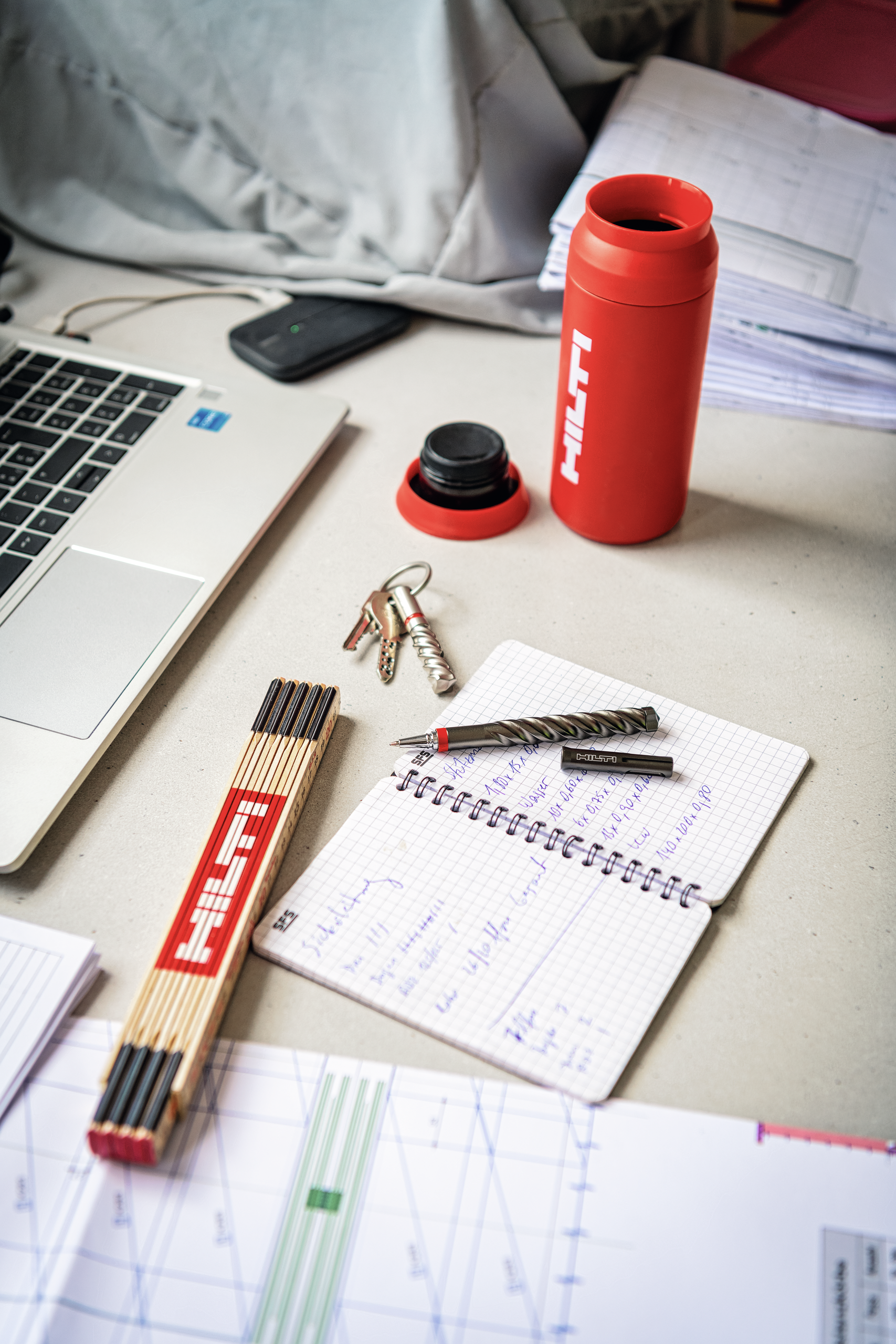 Workspace scene with a laptop, notebook, drill bit and pen, keys with a spiral anchor, a box of matches, and a red �������� bottle, suggesting engineering or construction-related note-taking and planning.