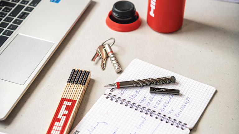 Workspace scene with a laptop, notebook, drill bit and pen, keys with a spiral anchor, a box of matches, and a red º£½ÇÉçÇø bottle, suggesting engineering or construction-related note-taking and planning.