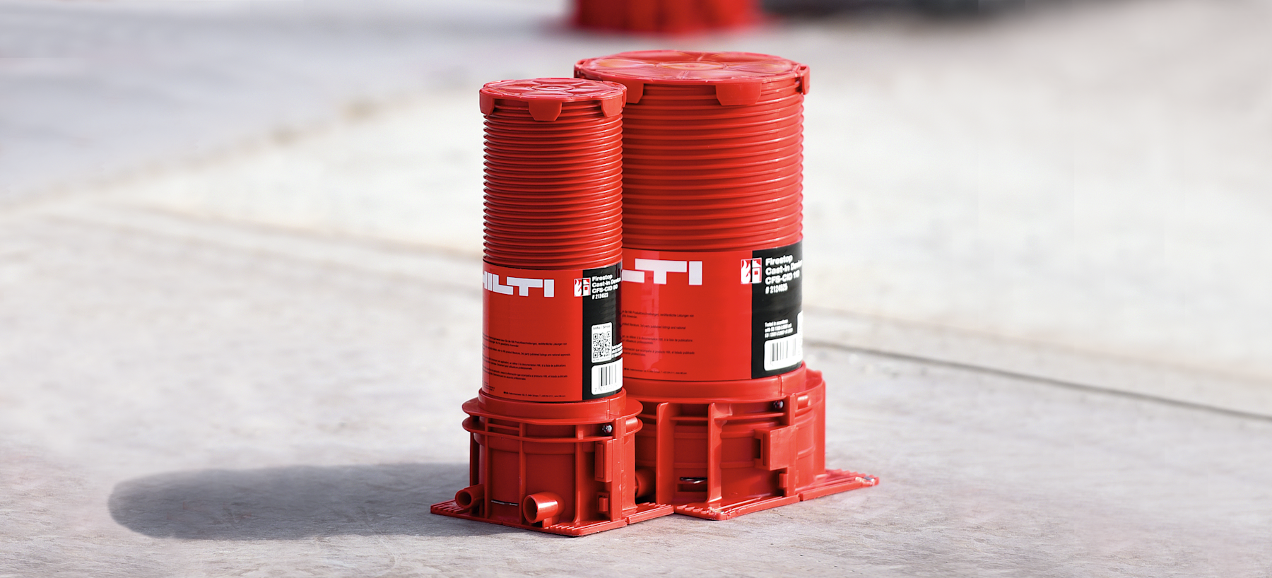 Two red cylindrical �������� components mounted on a base plate sit on a concrete surface, featuring ribbed sides and printed labels, likely used as protective or installation elements in construction.