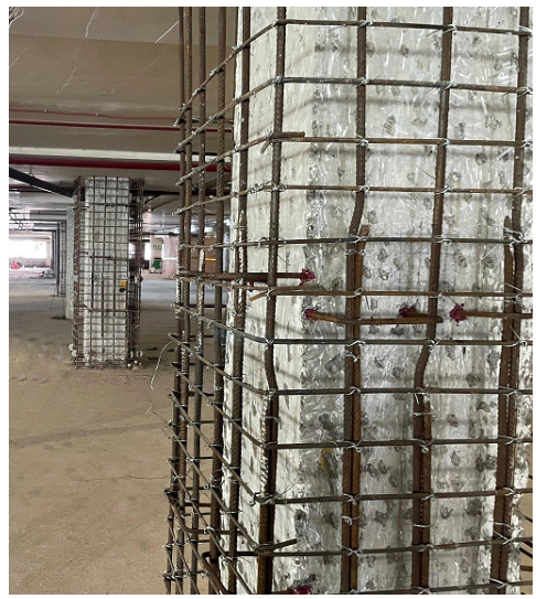 Concrete columns inside a building being strengthened with steel reinforcement cages. Multiple retrofitted columns are visible along the open floor space, demonstrating structural rehabilitation work.