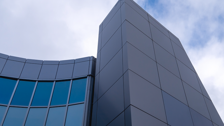 The corner of a grey coloured building made of metal composite panels and green reflective windows. Part of the building has a high tower and a lower curved section made of green windows.