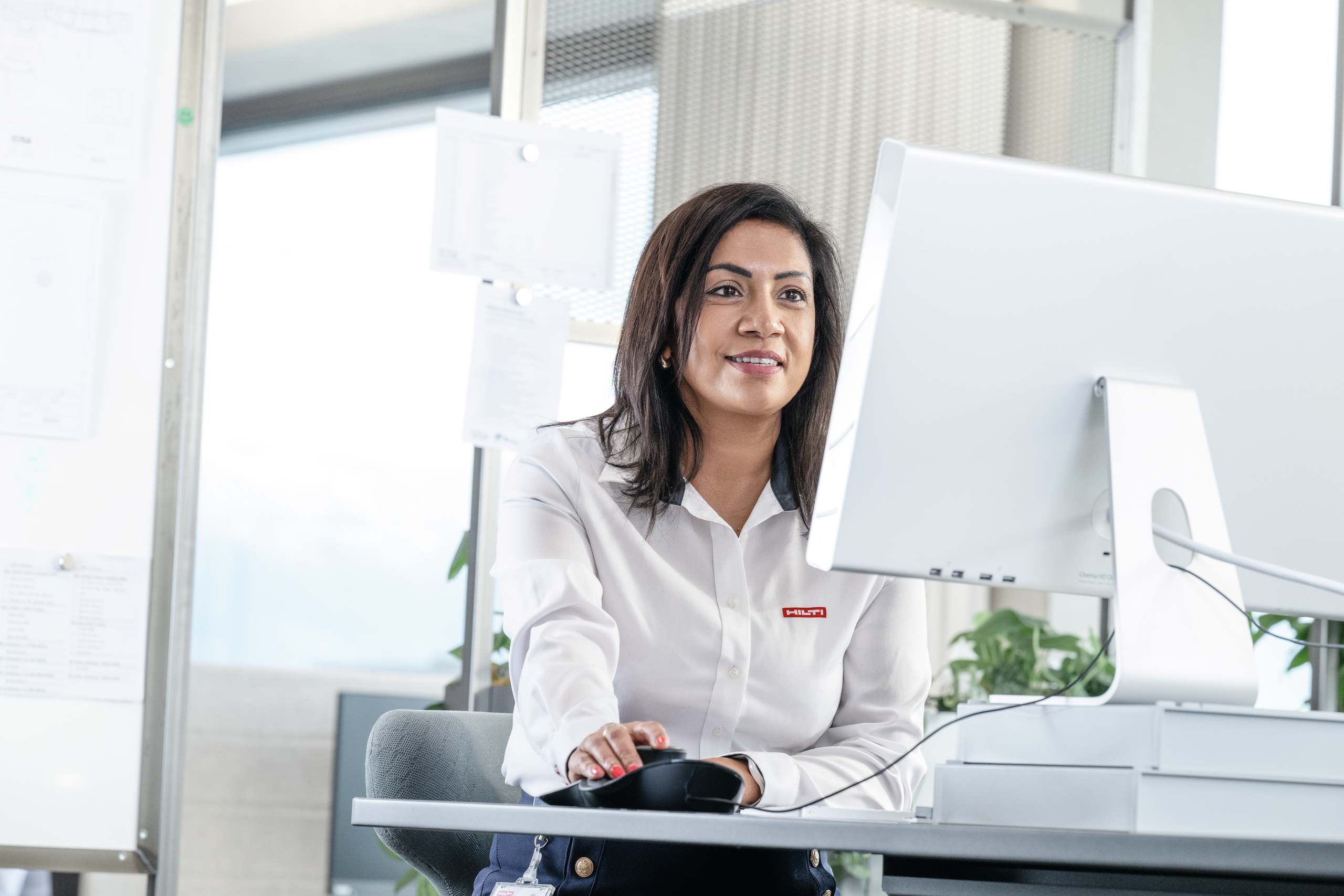 Office scene showing a person at a desk using a computer mouse and monitor, wearing a white shirt with a �������� logo, in a bright workspace with plants and office materials in the background.