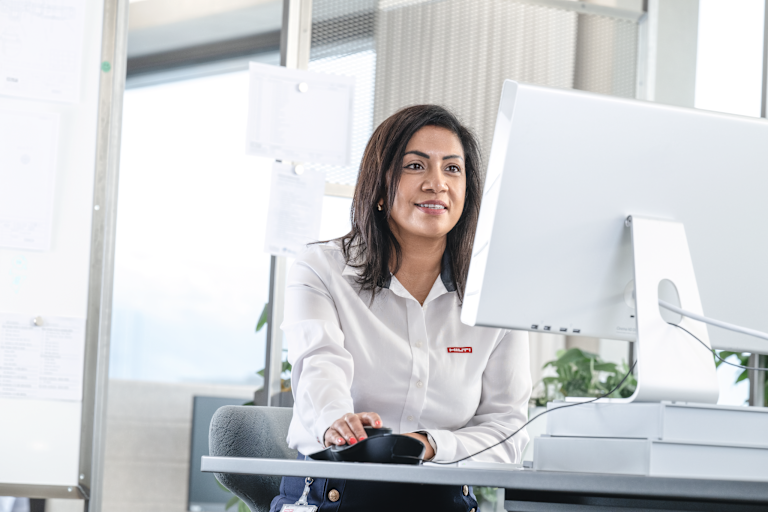 Office scene showing a person at a desk using a computer mouse and monitor, wearing a white shirt with a º£½ÇÉçÇø logo, in a bright workspace with plants and office materials in the background.