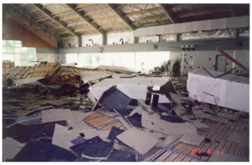 Damaged industrial building interior with collapsed roof materials and debris scattered across the floor, including broken panels, metal sheets, and insulation under a large, curved roof with exposed beams.