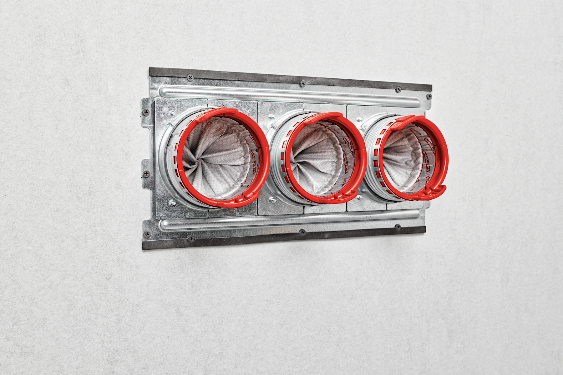 Metal firestop cable transit installed in a grey wall, featuring three circular cable openings with red retaining rings and sealed fabric membranes designed to maintain fire and smoke compartmentation.