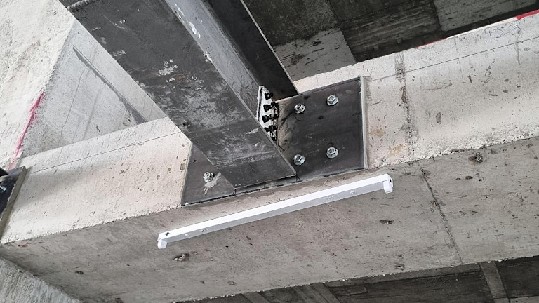 Steel beam connection with a baseplate anchored to a concrete soffit using multiple bolts, supporting a vertical steel member beneath a concrete structure.