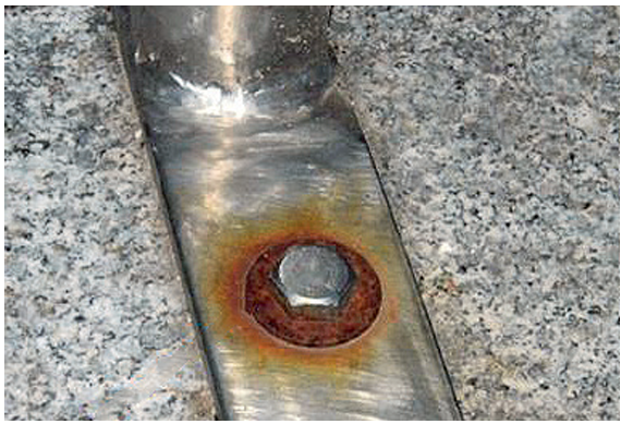 Close-up of a metal bolt in a plate with a ring of reddish rust around the base. The localized corrosion at the contact point suggests galvanic corrosion between dissimilar metals in an outdoor environment.