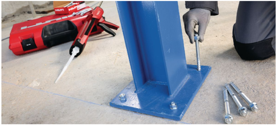 Photograph of a steel column base plate being anchored to a concrete floor. A hand positions an anchor rod beside the base plate, with additional anchors and a �������� adhesive dispenser visible in the background.