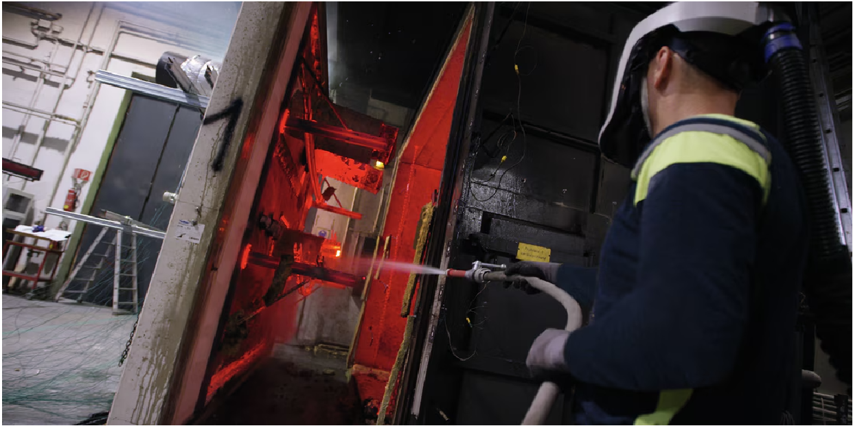 Person wearing protective equipment uses a high-pressure spray nozzle inside an industrial chamber glowing red-orange. Water mist is visible as the spray is directed at metal components during a testing or treatment process.