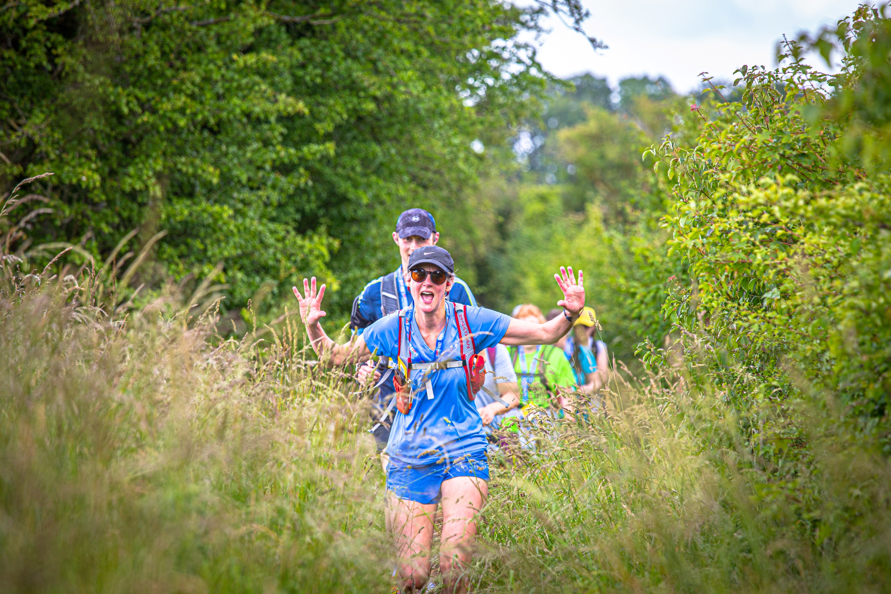 Four people in a line walking through an overgrown meadow. The person in front is wearing sunglasses and a cap and has their hands in the air.