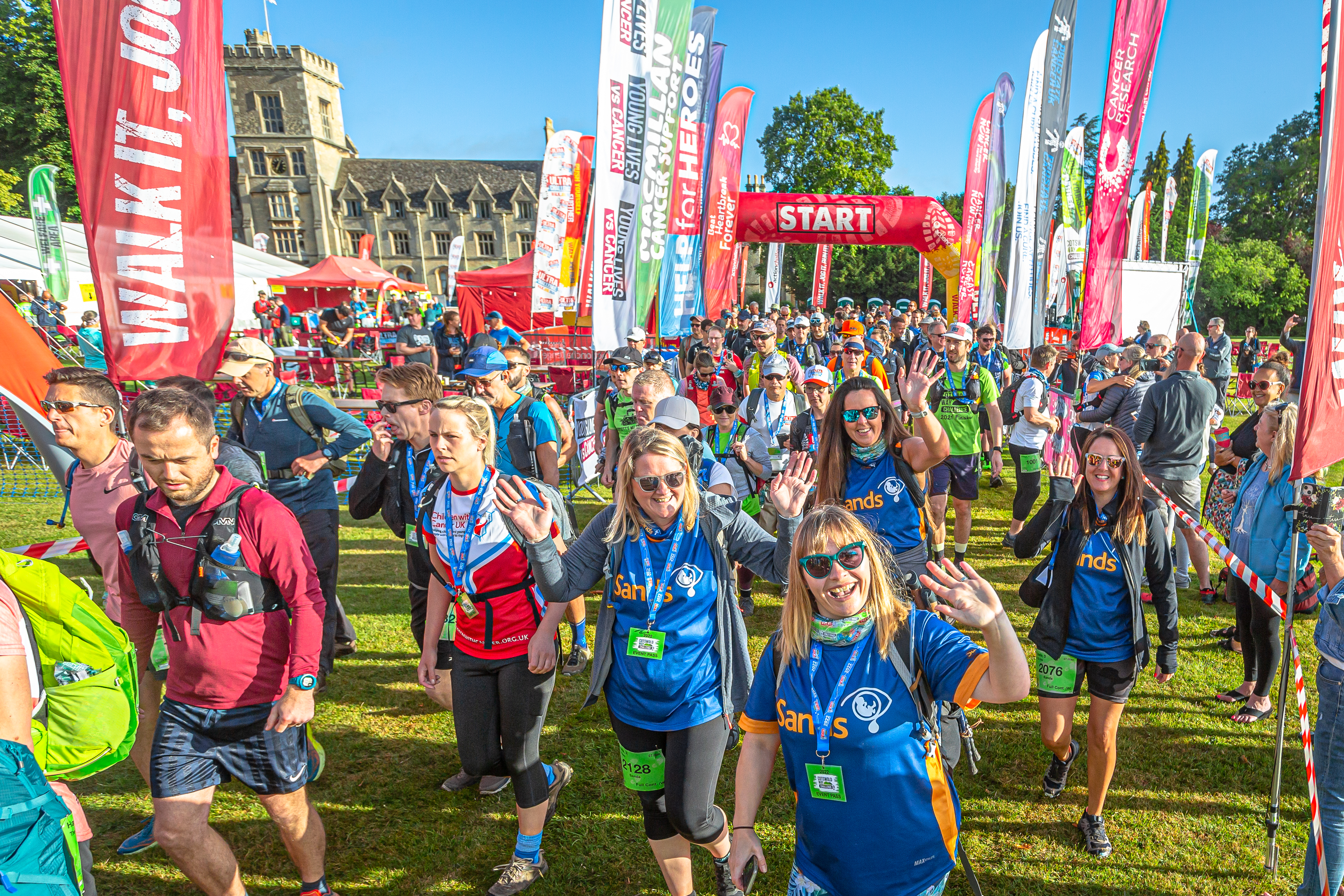 A large group of people smiling and waving as they start the Cotswold Way Ultra Challenge. Behind them is a large old building and red archway with 'Start' written on it.