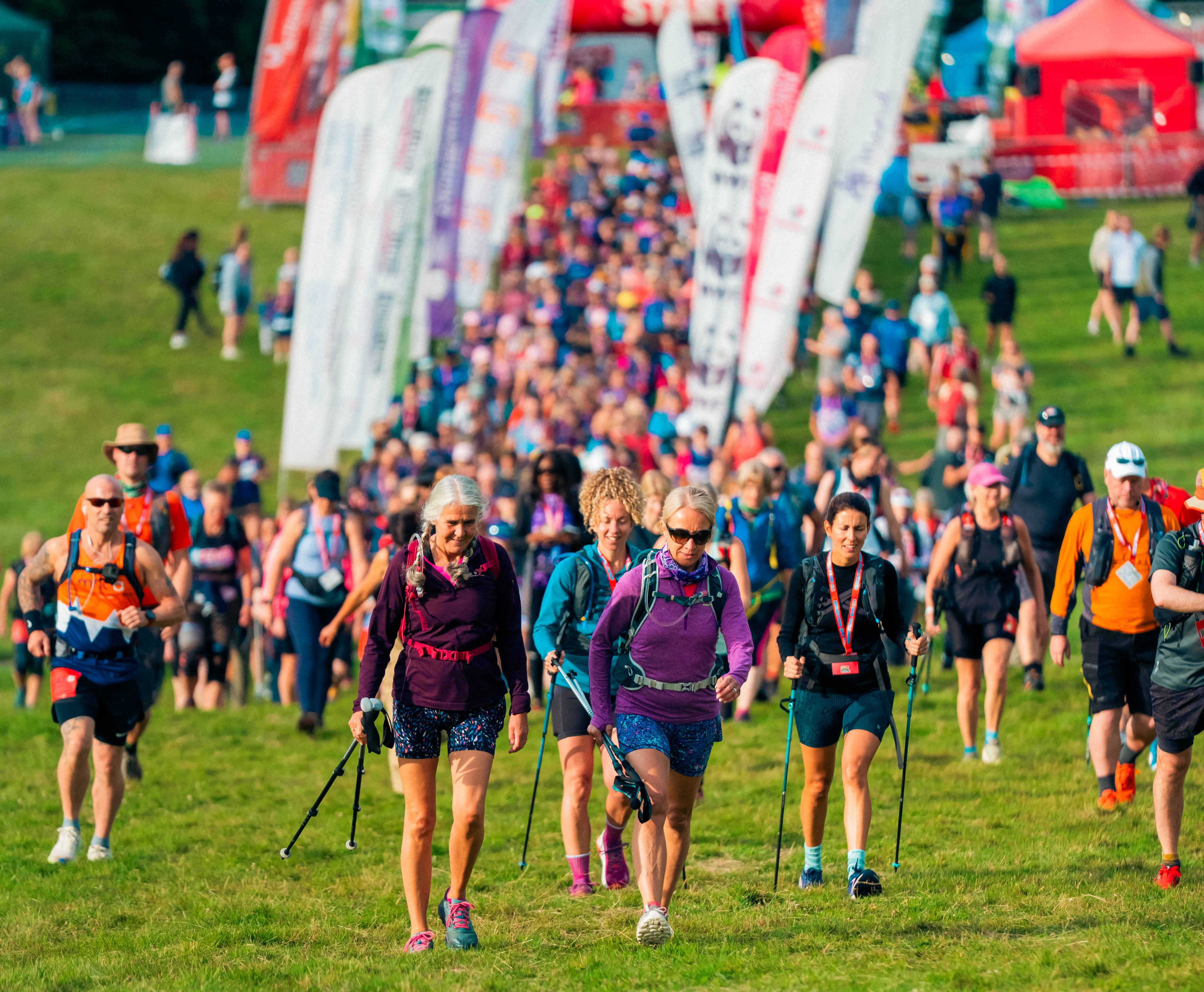 A large group of walkers, many with walking sticks and backpacks, walking across a field with lots of flags and a red archway with 'Start' in the background.