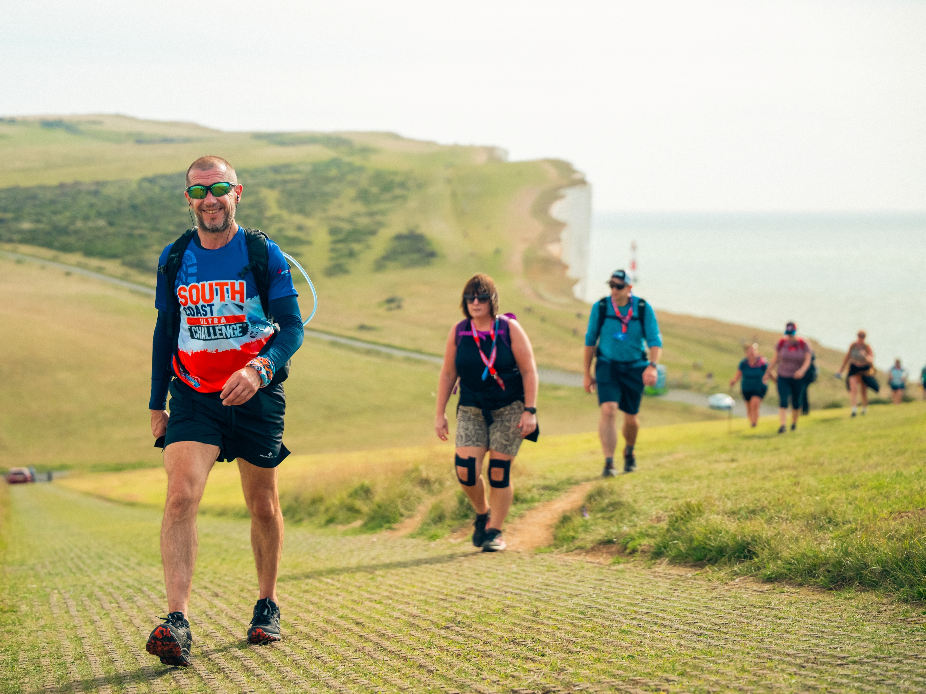 A group of people in a line walking along the South Downs coastal path. In the background is a road, white cliffs, and the sea.