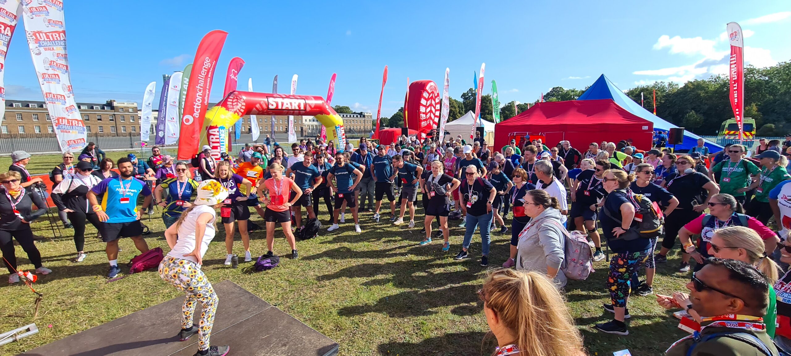 A large crowd of people outside all with their hands on their hips facing an instructor. There are lots of flags in the background, a large tent, and an archway saying 'Start' on it.