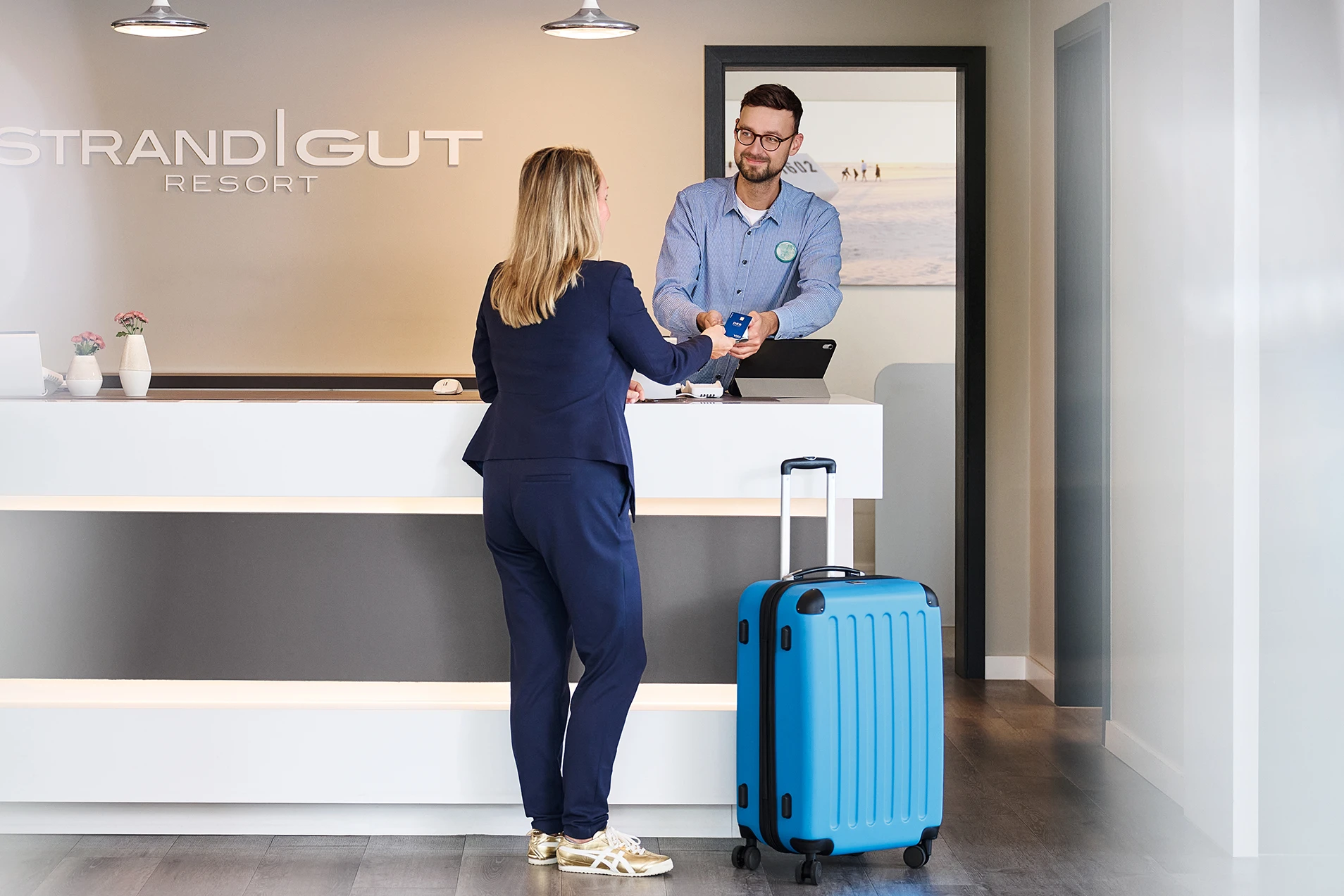Person checkt an der Rezeption des Strandgut Resorts mit blauem Koffer ein