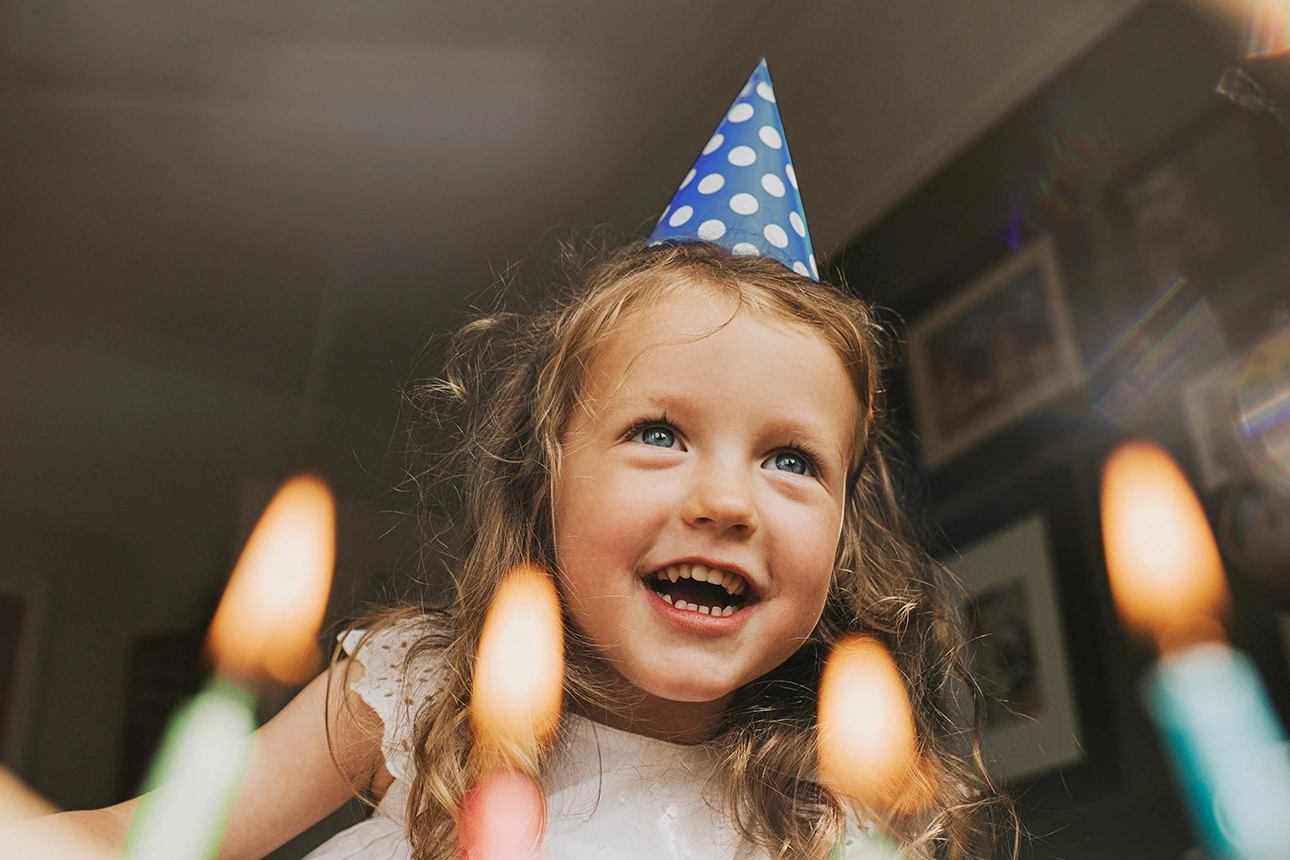 Mädchen sitzt vor einem Geburtstagskuchen und pustet Kerzen aus.