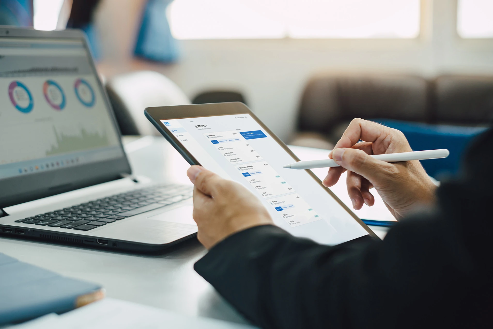 Person analysiert Finanzdaten auf einem Tablet neben einem Laptop im Büro