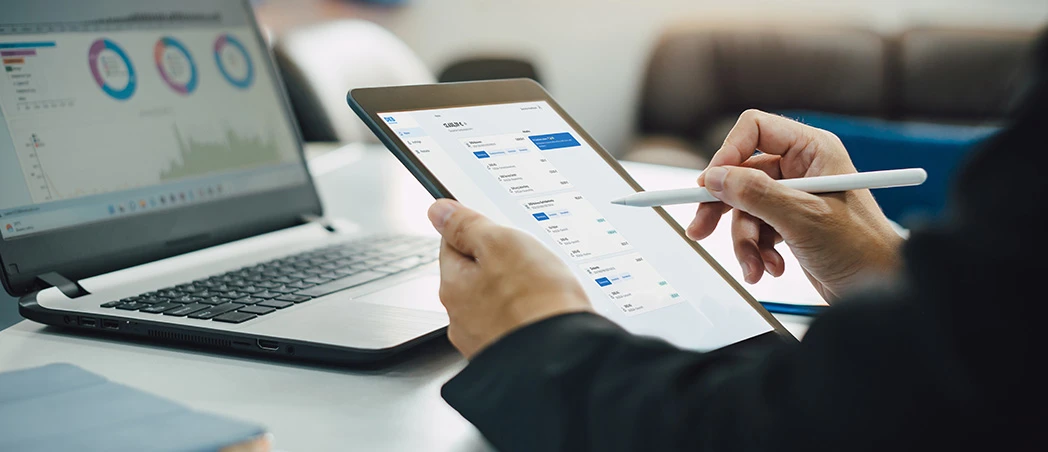 Person analysiert Finanzdaten auf einem Tablet neben einem Laptop im Büro