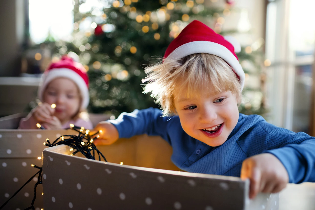 Zwei Kinder mit Weihnachtsmützen auf dem Kopf sitzen in Geschenkkisten. Im Hintergrund ist ein Weihnachtsbaum zu sehen.
