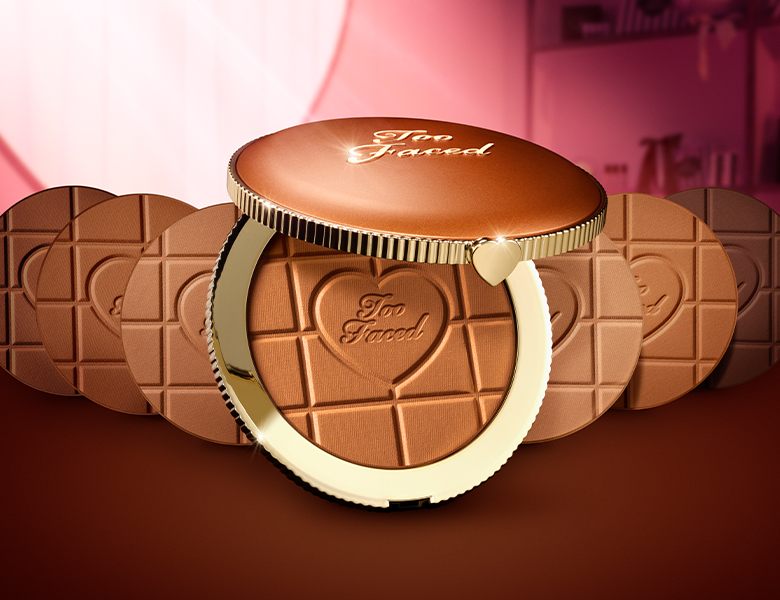 Her ses et billede med bronzere fra Too Faced placeret mod en pink baggrund. Produkterne er i runde kompakter med hjerteformede detaljer og teksten “Too Faced”.
