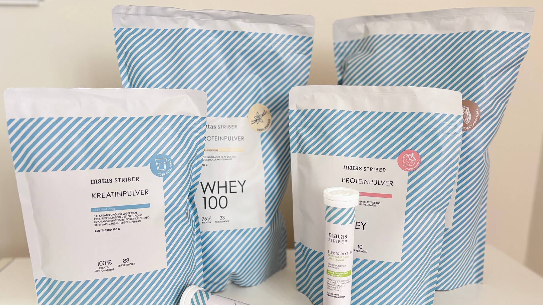 Flere produkter fra Matas Striber Sport står opstillet på en lys overflade foran en neutral baggrund. Produkterne består af store poser med kreatinpulver og proteinpulver i forskellige varianter samt et mindre rør med elektrolytter. Emballagerne er hvide med lyseblå diagonale striber og sort tekst. Produkterne står tæt samlet i forskellige højder, og nogle af poserne har små smagsikoner som vanilje, jordbær og chokolade på forsiden.