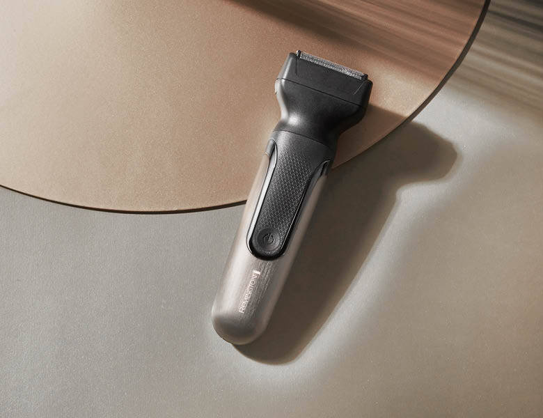 Her ses et billede af en elektrisk trimmer placeret på en lys overflade med en rund plade i baggrunden. Trimmeren er i grå og sort design med en tænd/sluk-knap på fronten.
