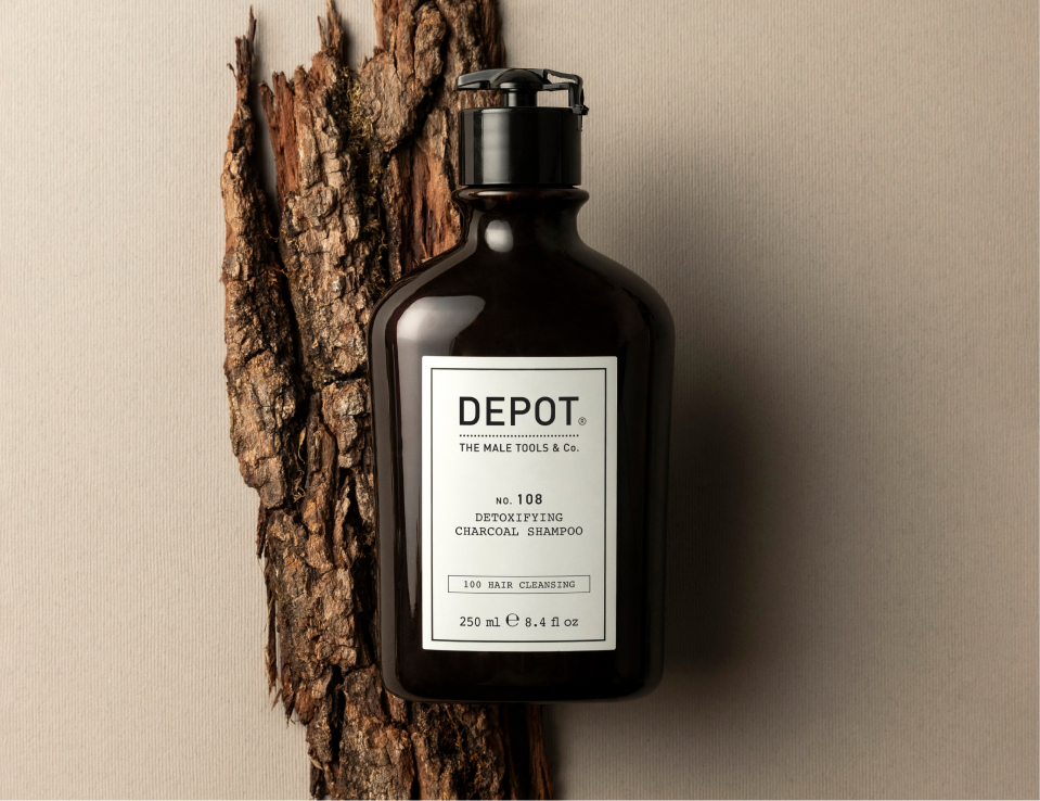 Her ses DEPOT No. 108 Detoxifying Charcoal Shampoo i en mørk flaske med sort pumpe, placeret foran et stykke træbark.
