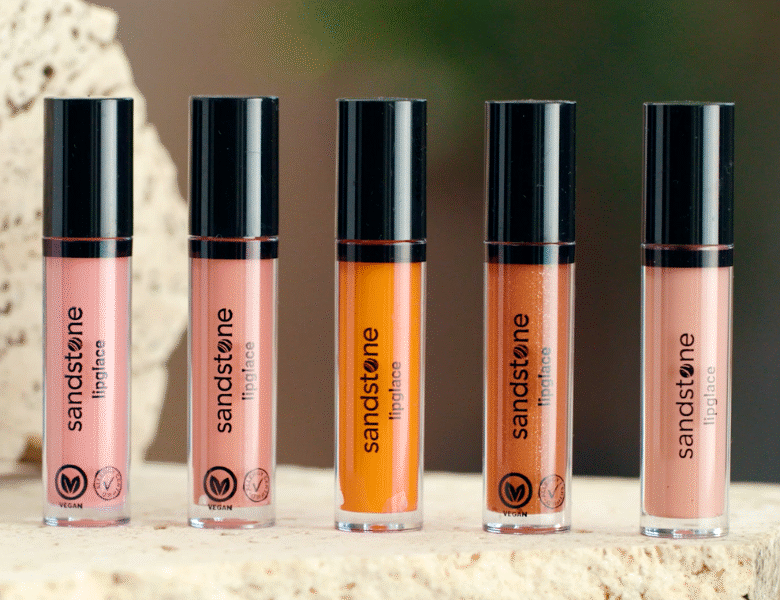 Her ses et billede med fem lipgloss-produkter fra Sandstone placeret side om side på en lys overflade. På produkterne står teksten “sandstone lipglace”, og de er i forskellige nuancer fra lyserød til orange og brun med sorte låg.
