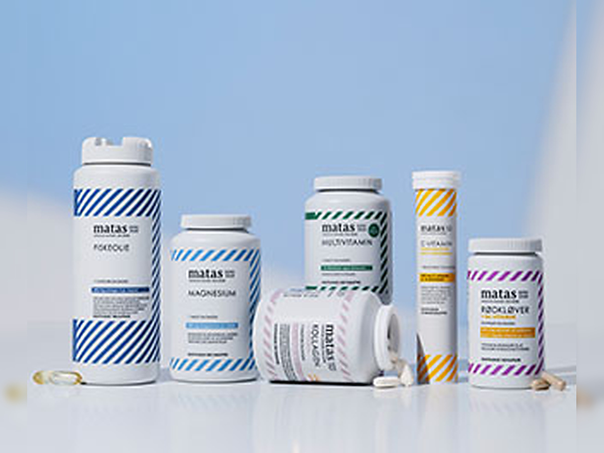 En hånd holder en stor bøtte Matas Striber fiskeolie ved siden af C-vitamin gummies og D-vitamin på lys baggrund.