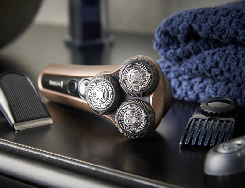 Her ses et billede af en elektrisk barbermaskine med tre roterende skær placeret på en overflade. Omkring den ligger forskellige tilbehørsdele og en blå klud i baggrunden.