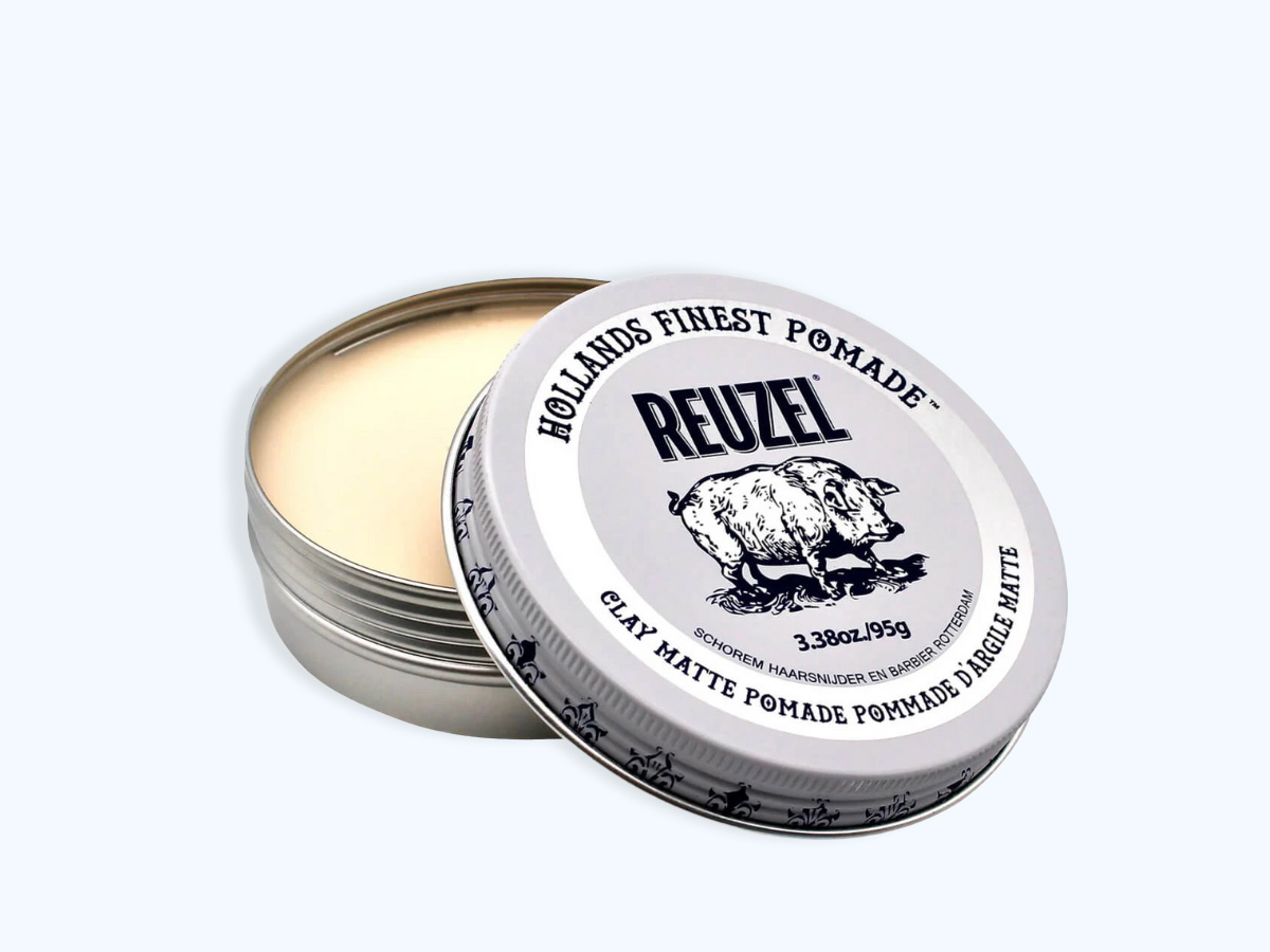 Her ses et billede af en åben dåse Reuzel Clay Matte Pomade mod en lys baggrund. Låget er placeret skråt oven på dåsen, så både produktets lyse voks og etiketten med Reuzel-logoet og det karakteristiske vildsvin er synlige. På emballagen står blandt andet “Holland's Finest Pomade” og “Clay Matte Pomade”.