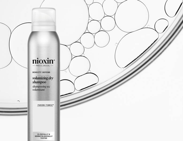 Her ses et billede af en dry shampoo fra Nioxin placeret mod en lys baggrund med grafiske boblemønstre. På produktet står teksten “nioxin PRO CLINICAL volumizing dry shampoo”. Flasken er i sølvfarvet design.