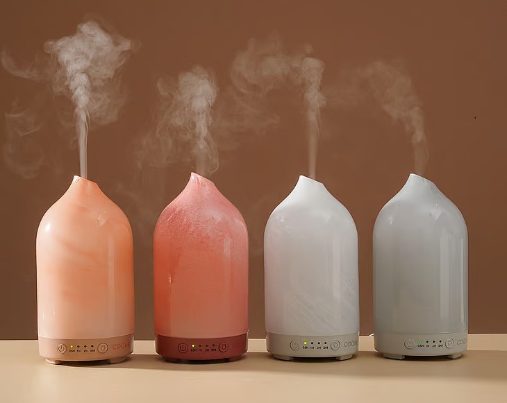 Fire Coohe aromadiffusere i rosa og hvide nuancer, der udsender damp, opstillet på række mod en varm brun baggrund.