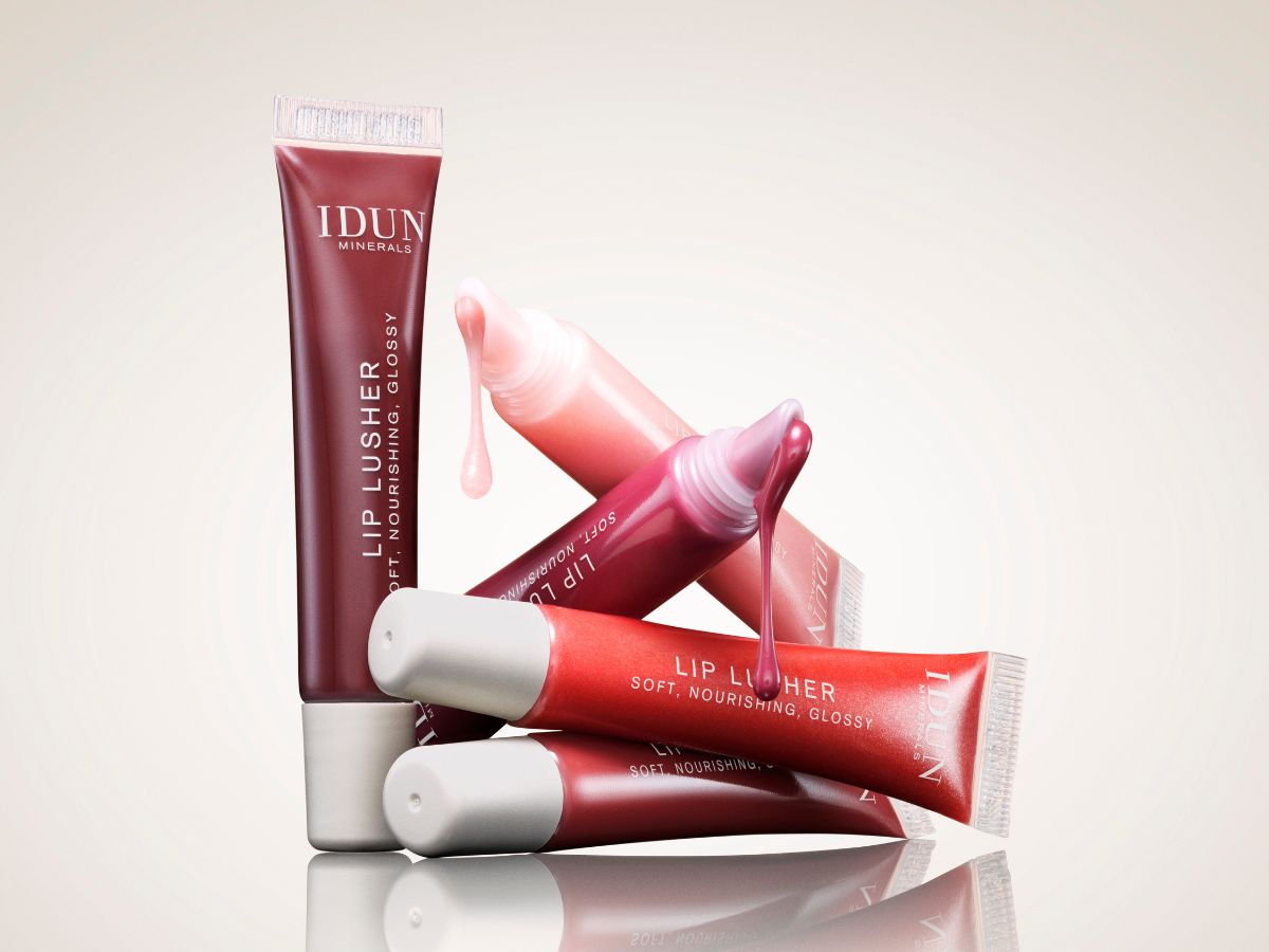 Her ses et billede med flere Lip Lusher lipgloss fra IDUN Minerals mod en lys baggrund. Lipglossene er arrangeret oven på hinanden i forskellige rosa og røde nuancer, og nogle af tuberne er åbne, så den glansfulde tekstur løber ud af applikatoren. Billedet fremhæver produkternes farver og blanke finish.