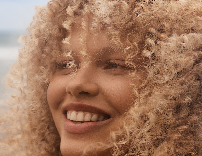 Her ses et nærbillede af en smilende person med lyst, tæt krøllet hår, fotograferet udendørs.