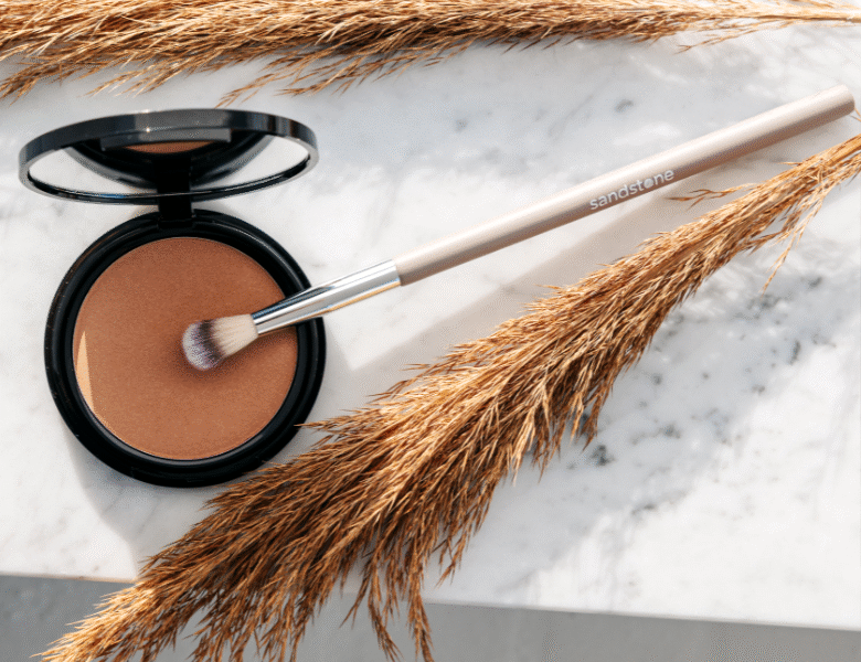 Her ses et billede af en pudder og en makeupbørste fra Sandstone placeret på en marmorlignende overflade. På børsten står teksten “sandstone”. Produktet er i en varm brun nuance og omgivet af tørrede planter.