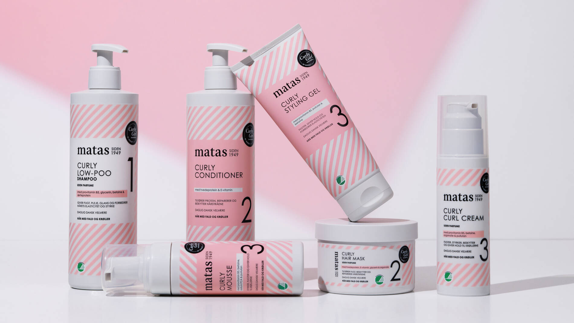 Et udvalg af Matas Curly-hårprodukter står opstillet på en hvid overflade foran en lyserød baggrund. Produkterne har hvide emballager med lyserøde striber og består blandt andet af shampoo, conditioner, stylinggel, curl cream, mousse og hårmaske. Nogle produkter står oprejst med pumpe, mens en mousseflaske ligger ned foran produkterne. En tube med stylinggel balancerer skråt oven på en rund krukke med hårmaske. Flere af produkterne har det grønne Svanemærke nederst på emballagen.