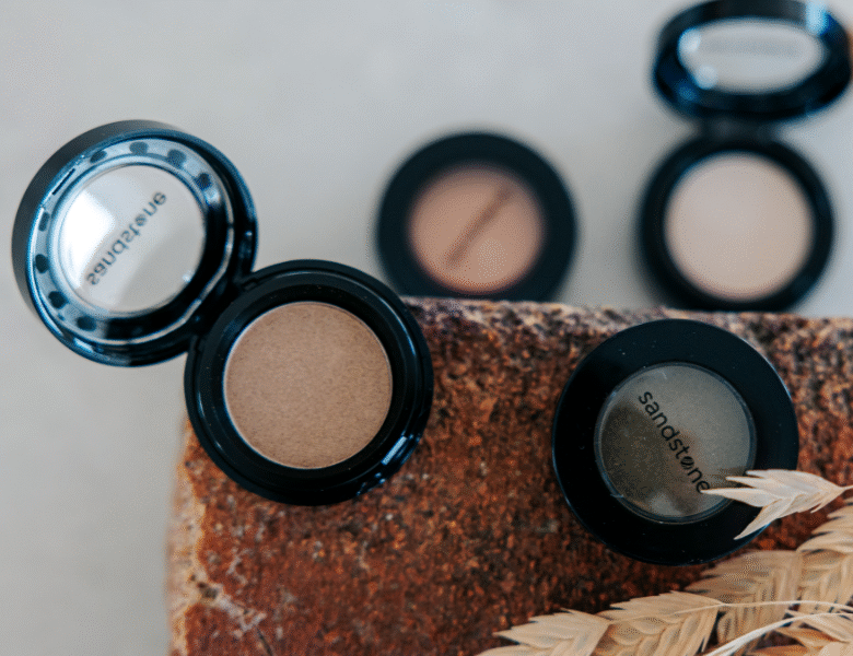 Her ses et billede med flere øjenskygger fra Sandstone placeret på en overflade. Nogle af produkterne er åbne, og på dem står teksten “sandstone”. Farverne er i neutrale nuancer som brun og beige.