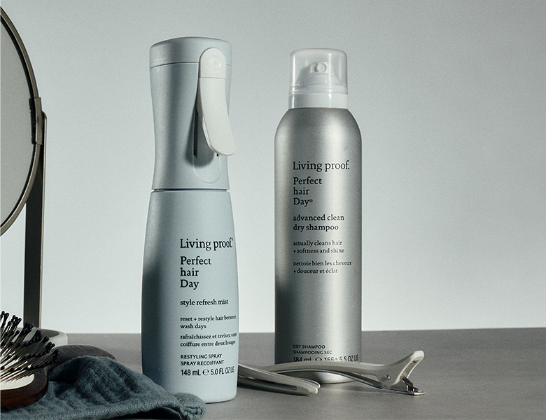 Her ses Living Proof Perfect Hair Day Style Refresh Mist og Perfect Hair Day Advanced Clean Dry Shampoo stående sammen med en hårbørste, hårklemmer og et spejl mod en lys baggrund.